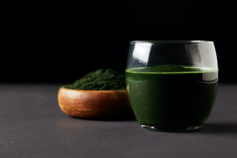 close up view of fresh spirulina drink in glass and spirulina powder in wooden bowl on black