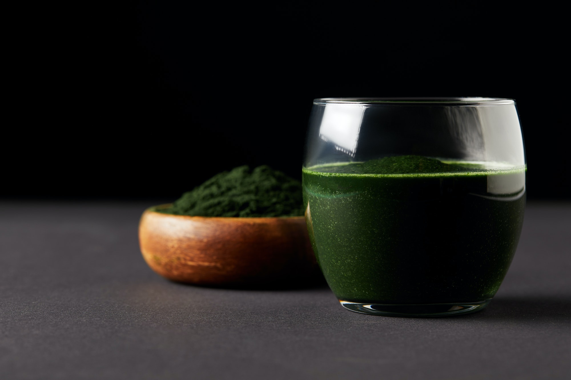 close up view of fresh spirulina drink in glass and spirulina powder in wooden bowl on black