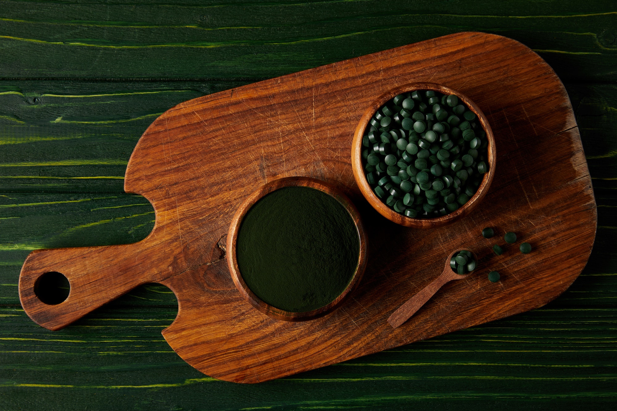 top view of cutting board with wooden spoon, bowls with spirulina powder and spirulina in pills on